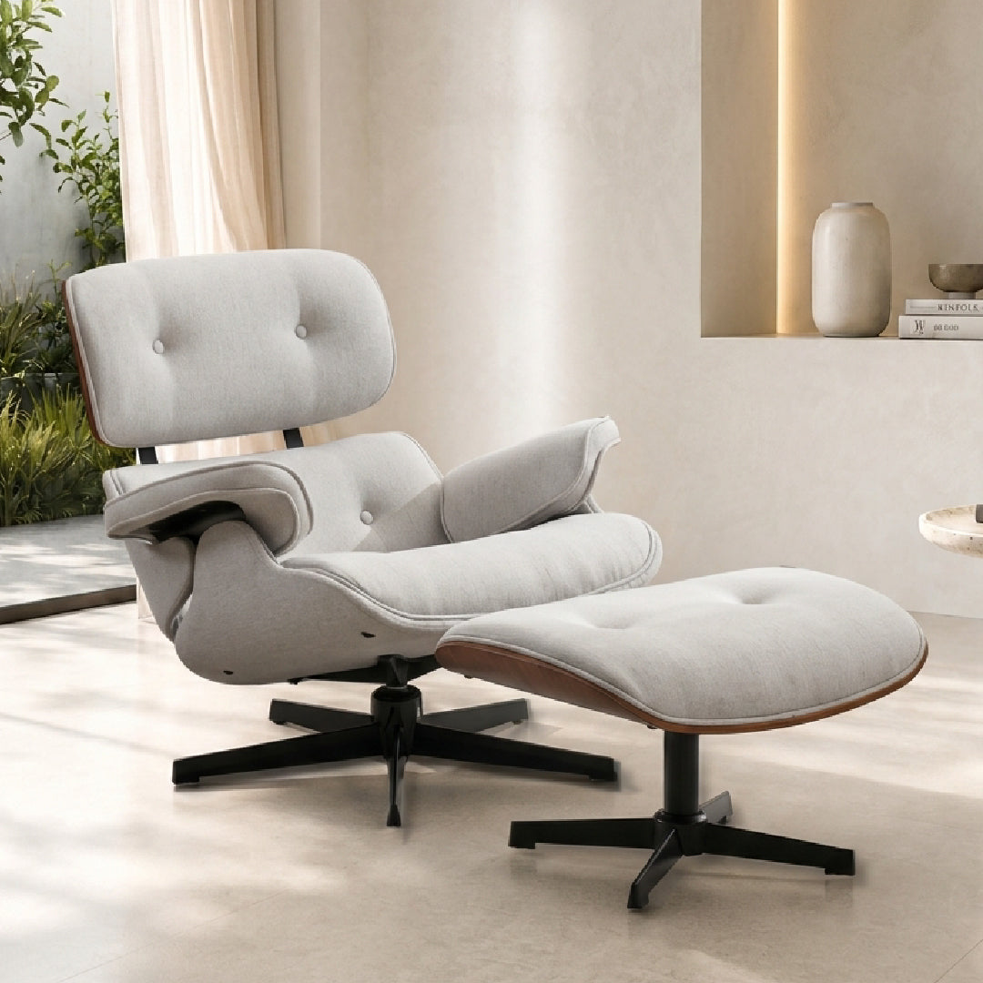 A modern lounge chair with matching ottoman in light beige fabric, shown in a bright, minimalist interior setting with warm natural light.