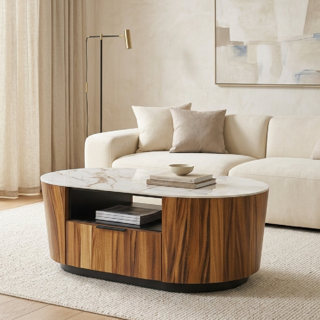 An oval coffee table with a warm wood grain finish and a white marble top, styled in a modern living room setting with a cream sofa and neutral decor.
