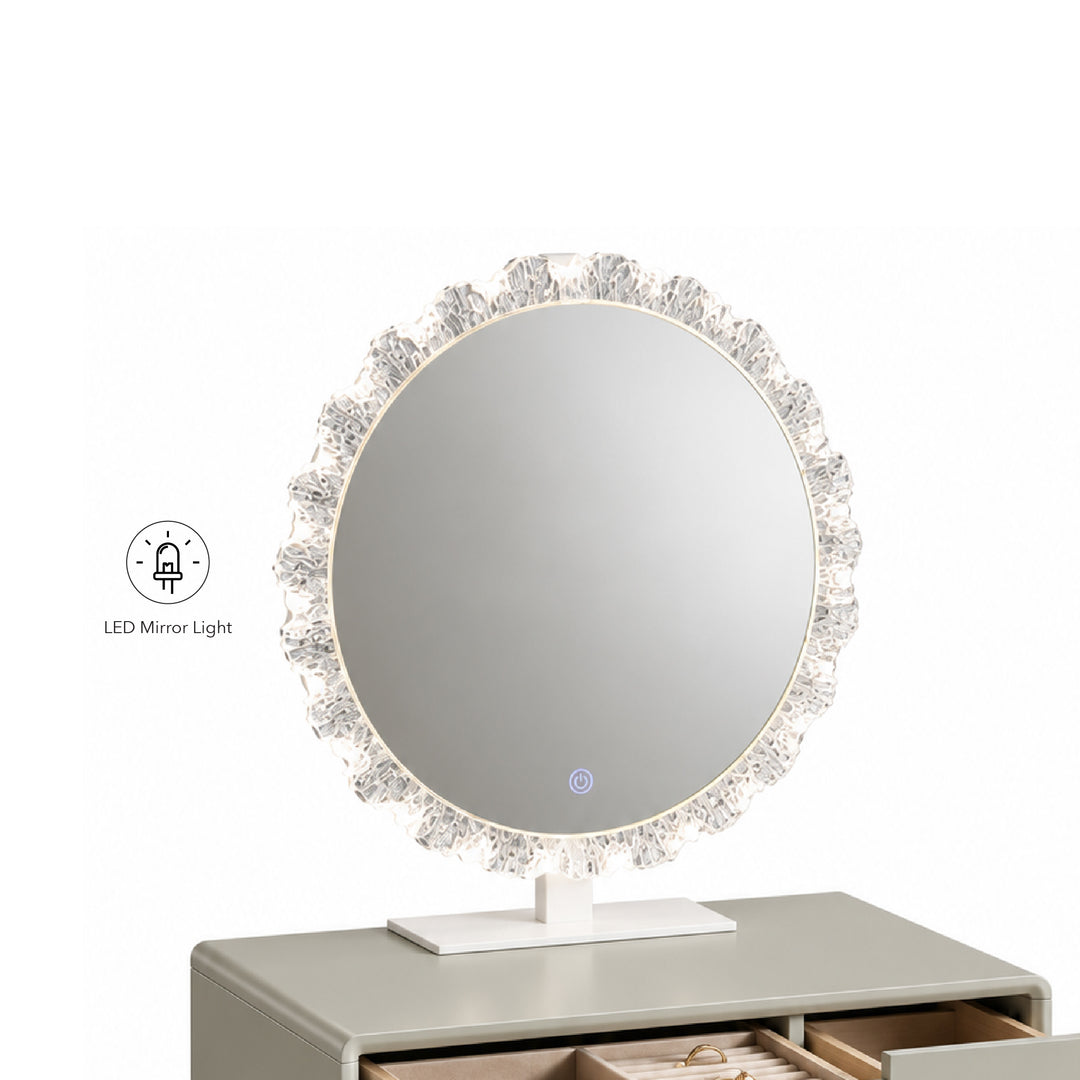 Round LED vanity mirror with a crystal-embellished frame and white stand, shown on a gray dresser against a white background. A small icon in the lower left indicates LED mirror light.