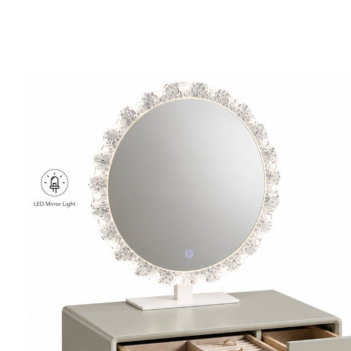 Round LED vanity mirror with a crystal-embellished frame and white stand, shown on a gray dresser against a white background. A small icon in the lower left indicates LED mirror light.