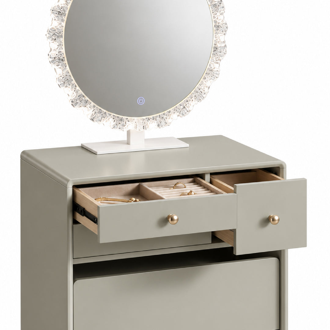 A modern makeup vanity table in a light gray finish with two open drawers displaying jewelry, a large round mirror with a crystal-like frame, and gold-tone knobs, set against a plain white background.