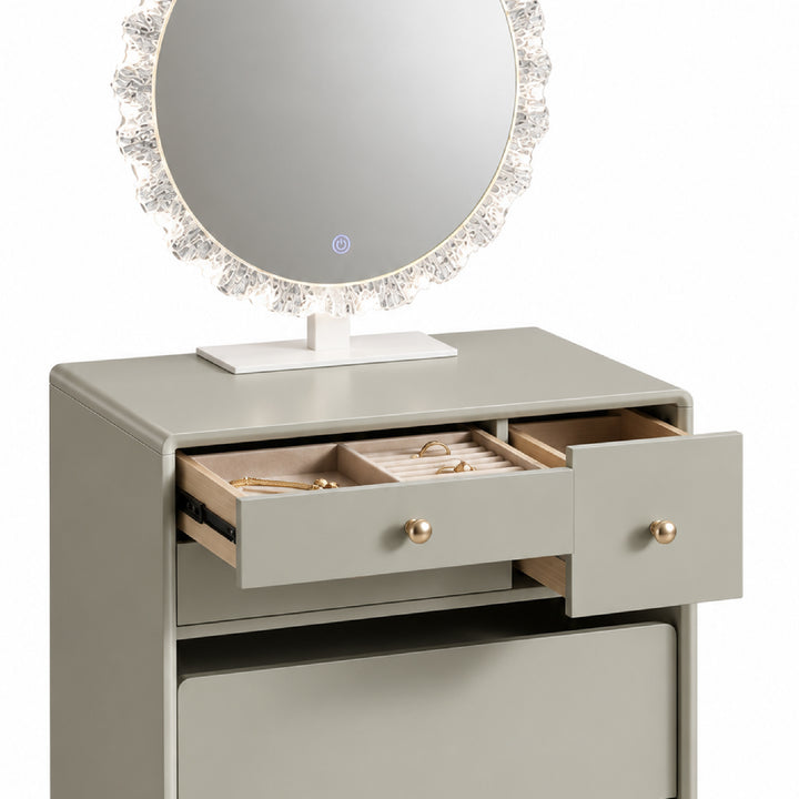 A modern makeup vanity table in a light gray finish with two open drawers displaying jewelry, a large round mirror with a crystal-like frame, and gold-tone knobs, set against a plain white background.