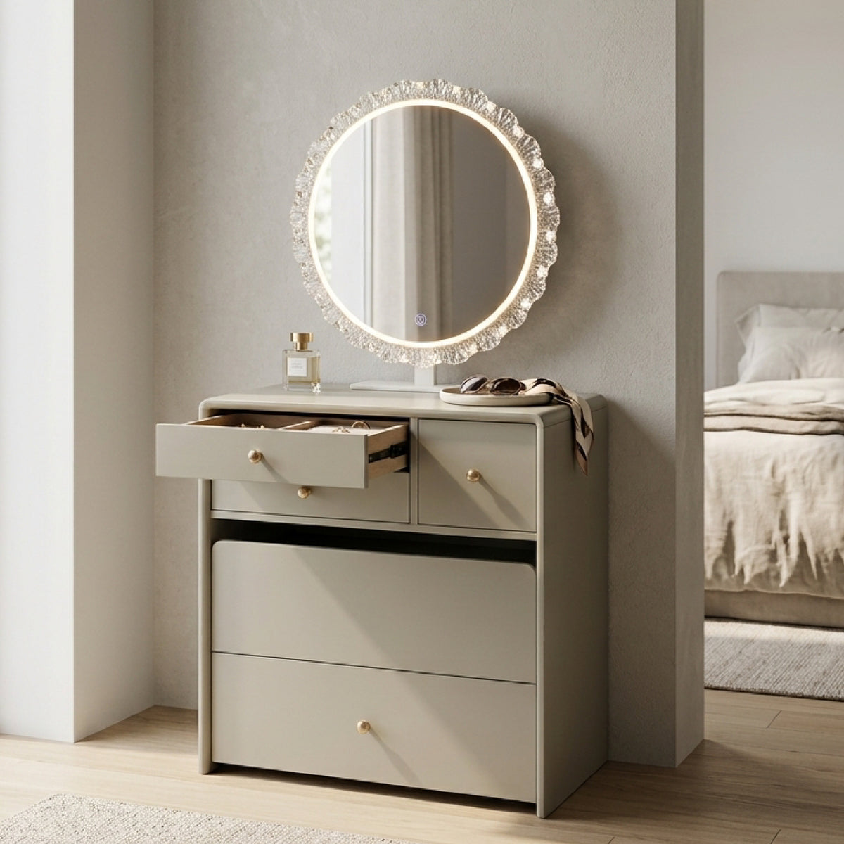 A modern light beige vanity dresser with three drawers, one open, featuring a round illuminated mirror with a crystal-like frame. The dresser is styled with a perfume bottle and sunglasses in a bedroom setting.