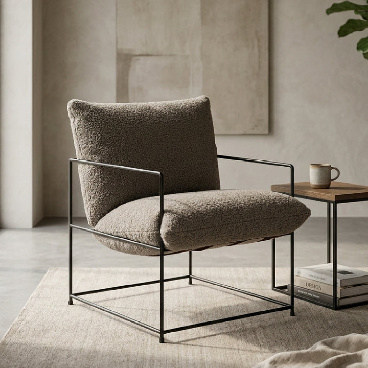 A modern armchair with a textured taupe bouclé seat and back cushion, supported by a black metal frame, shown in a styled living room setting next to a wooden side table.