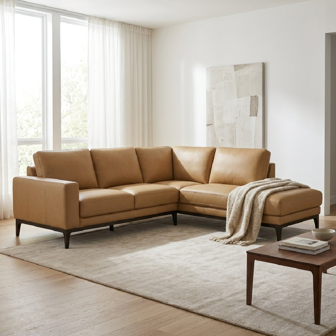 A tan leather sectional sofa with dark wooden legs, styled in a bright, modern living room with a light beige rug, a small wooden coffee table, and a draped throw blanket.
