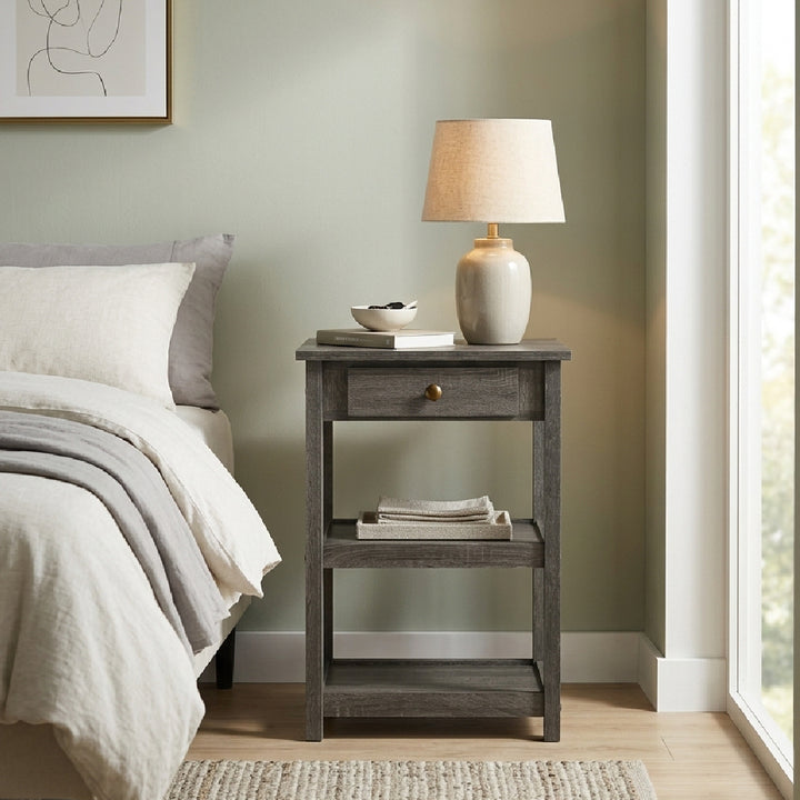 A gray wooden nightstand with a single drawer and open lower shelf, styled beside a bed in a softly lit bedroom. A beige table lamp, small bowl, and books sit on top, with folded linens on the shelf below.