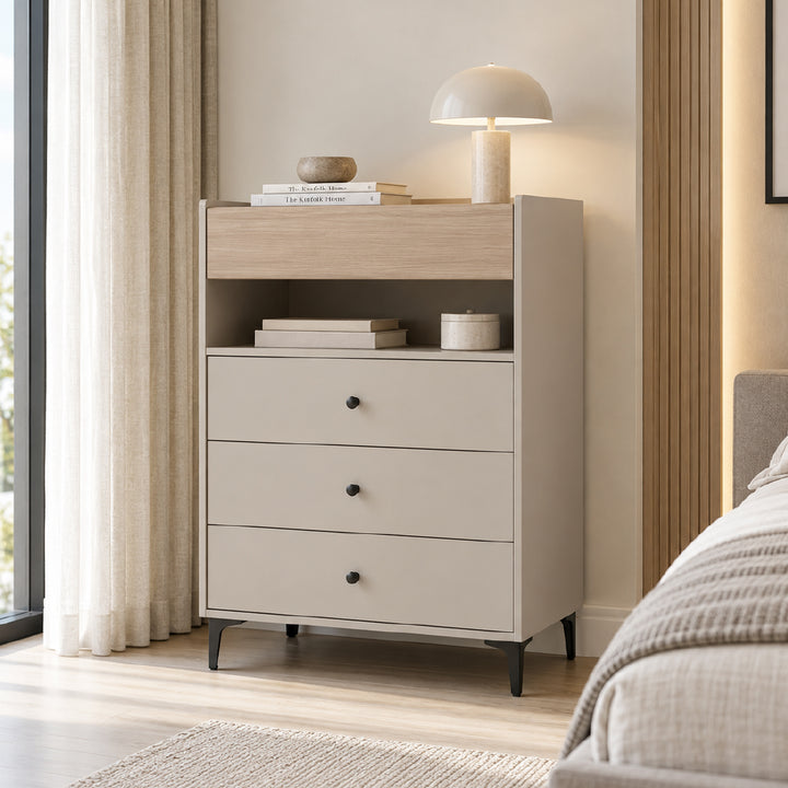 A three-drawer nightstand or storage cabinet in a light warm gray finish with a light wood top shelf, styled in a modern bedroom next to a bed and window.