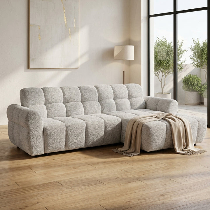 A light gray bouclé sectional sofa with a tufted backrest and chaise lounge, styled in a modern living room with a beige throw blanket, wood floors, and a large window.