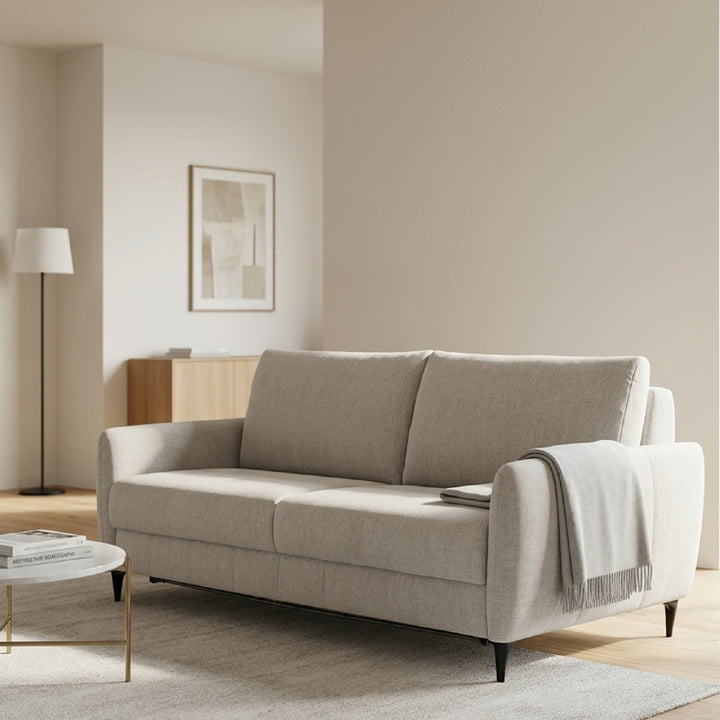 A light khaki upholstered loveseat sofa-bed in a modern, minimalist living room setting with a white throw blanket draped over one arm, a small round coffee table, and a floor lamp in the background.