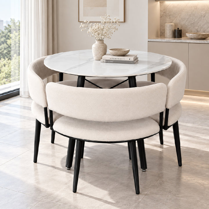 A modern round dining table with a white marble-look top and black legs, surrounded by four upholstered dining chairs in a warm off-white fabric with black legs, set in a bright, contemporary dining room.