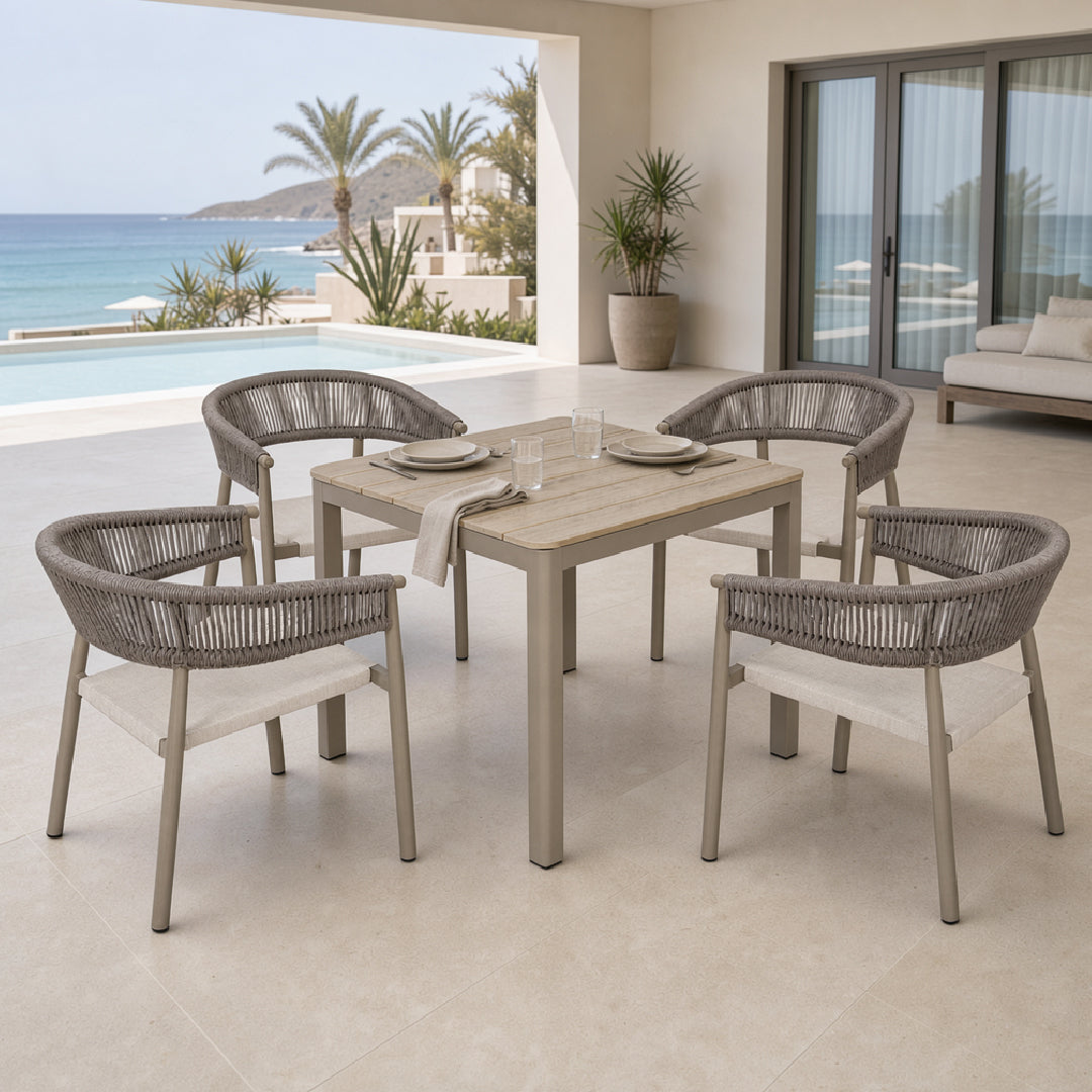 A 5-piece outdoor dining set featuring a rectangular light wood table and four woven gray armchairs with light beige seat cushions, set on a tiled patio with an ocean view background.