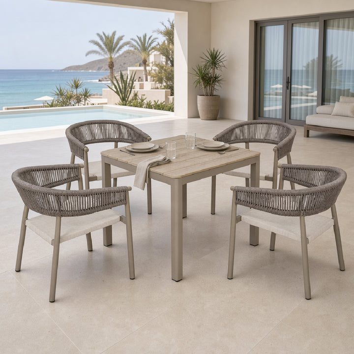 A 5-piece outdoor dining set featuring a rectangular light wood table and four woven gray armchairs with light beige seat cushions, set on a tiled patio with an ocean view background.