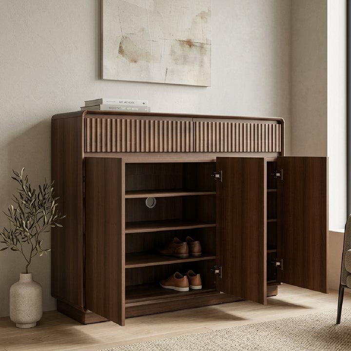 A dark brown wooden shoe cabinet with open doors, showing three shelves and shoes inside, styled in a modern minimalist room next to a vase with greenery.